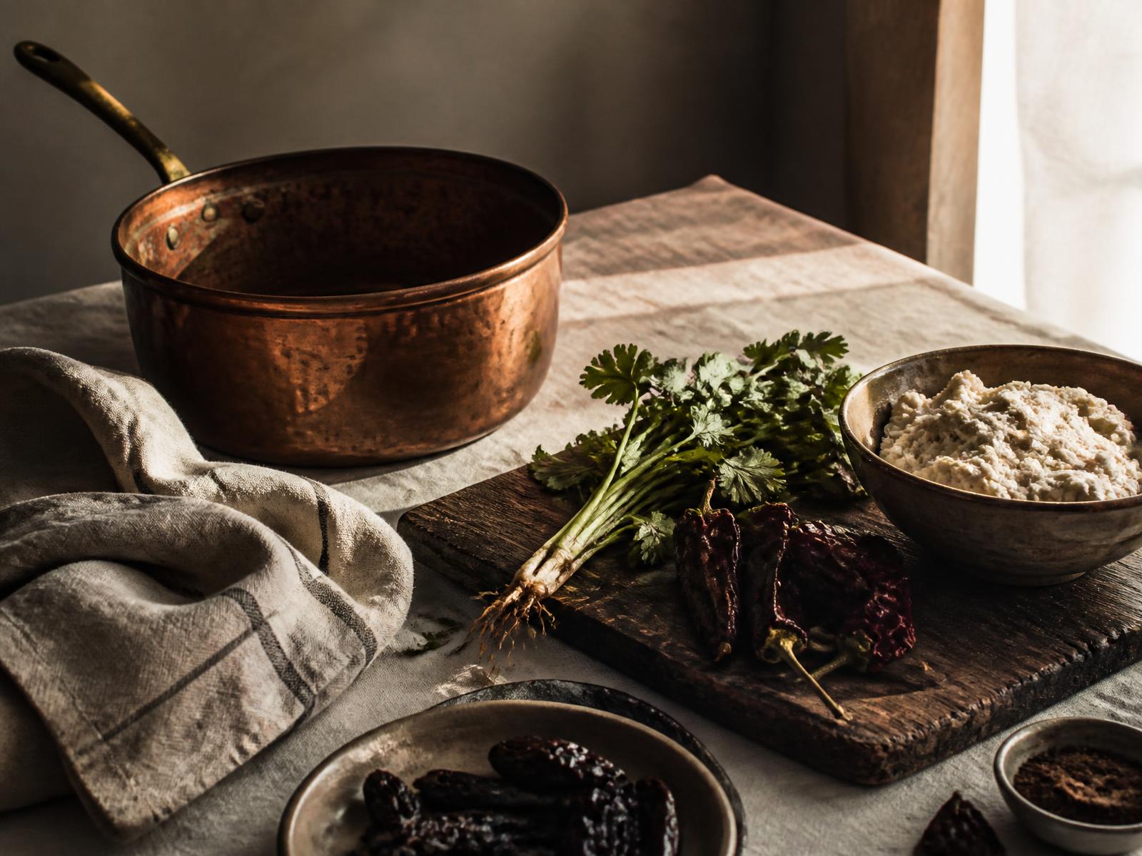 A copper pan, herbs, dried chiles and masa on a linen-covered kitchen table in soft natural light